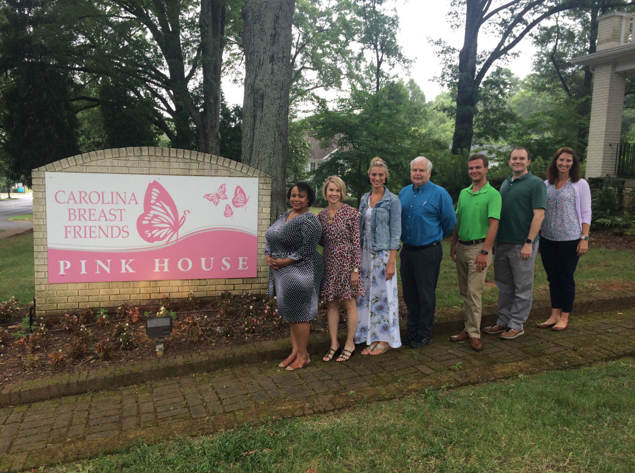 A team of people from Apparo, Rodgers Builders and Carolina Breast Friends posing by the Carolina Breast Friends sign in front of The Pink House.