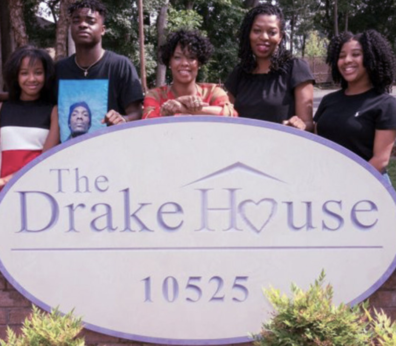Women and men standing outside behind a large sign that says The Drake House