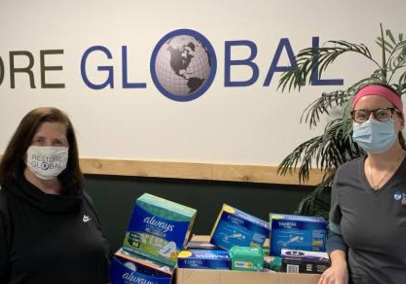 Two women wearing face masks with a delivery of office supplies in front of a Restore Global banner